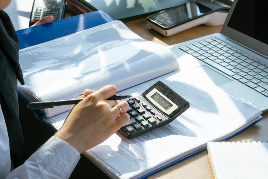 A person working on financial calculations using a calculator and laptop at an office desk.