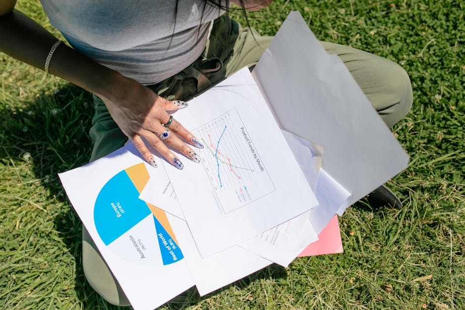 A woman sitting on grass analyzing business documents with charts and graphs in sunlight.